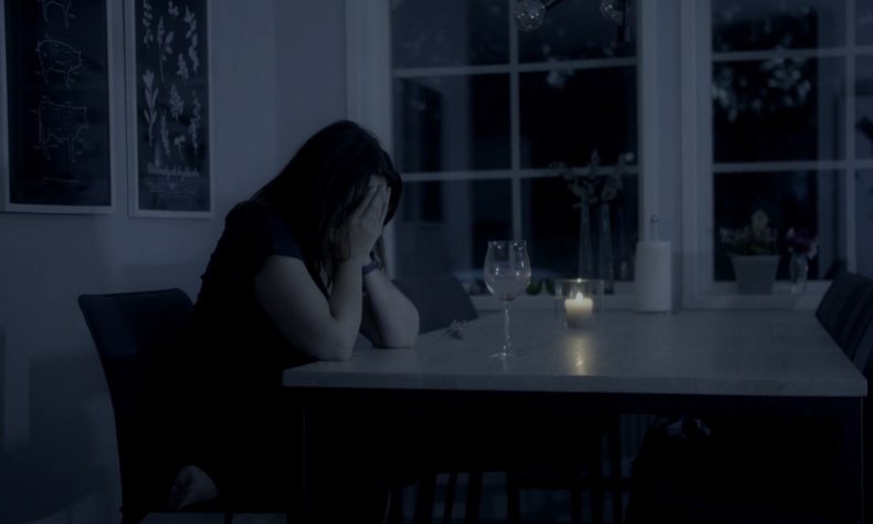 Žena sedi za stolom u zamračenoj prostoriji, drži lice rukama